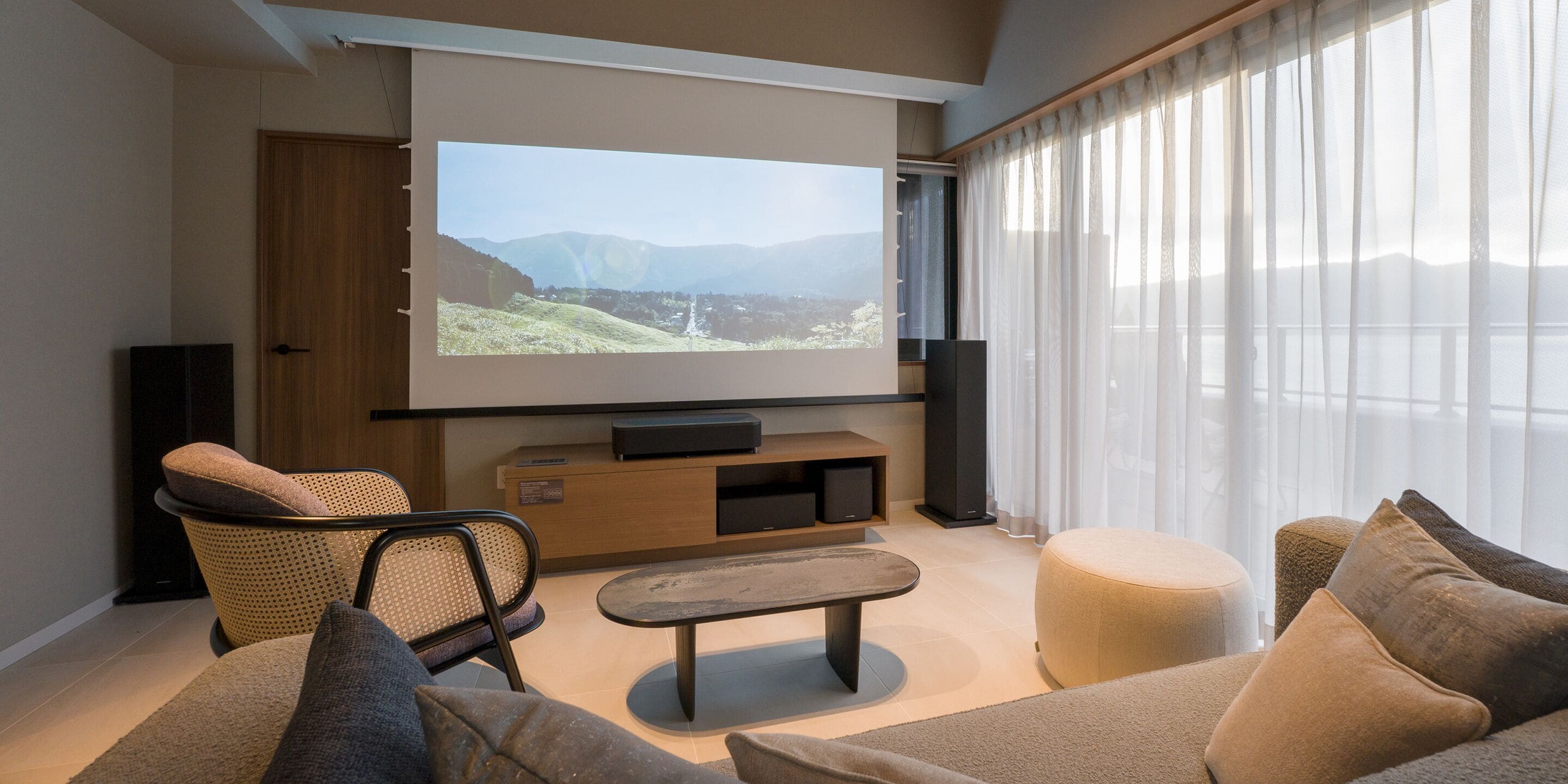 A modern living room with a large flat-screen TV mounted on the wall, a comfortable sofa, a small coffee table, and a view of a mountainous landscape through sheer curtains.