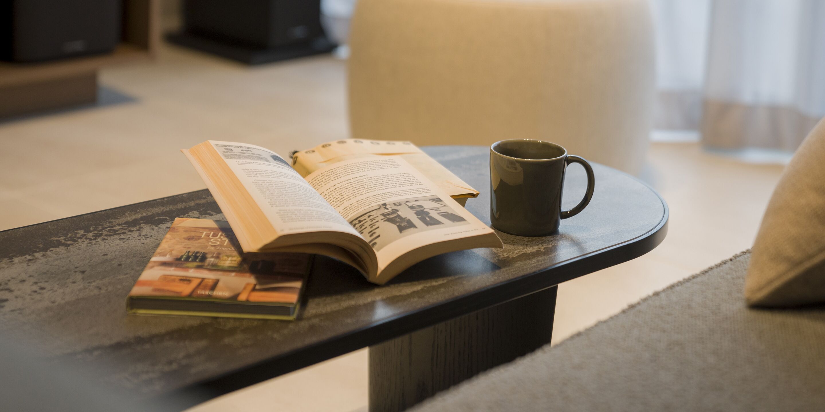 Ein Buch und eine Tasse auf einem Tisch, im Hintergrund ein Kissen.