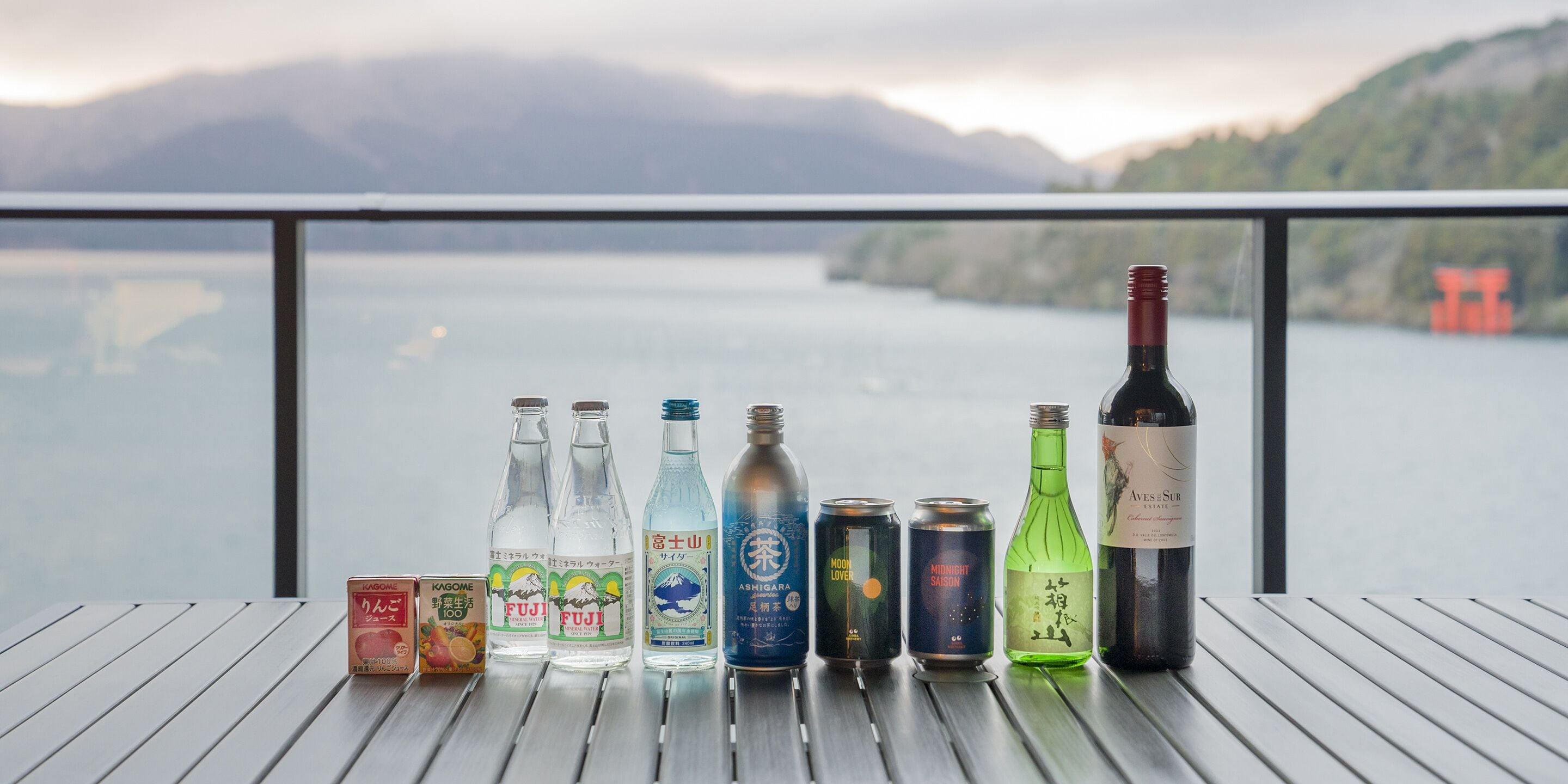 A variety of beverages on a wooden table with a scenic lake view in the background.
