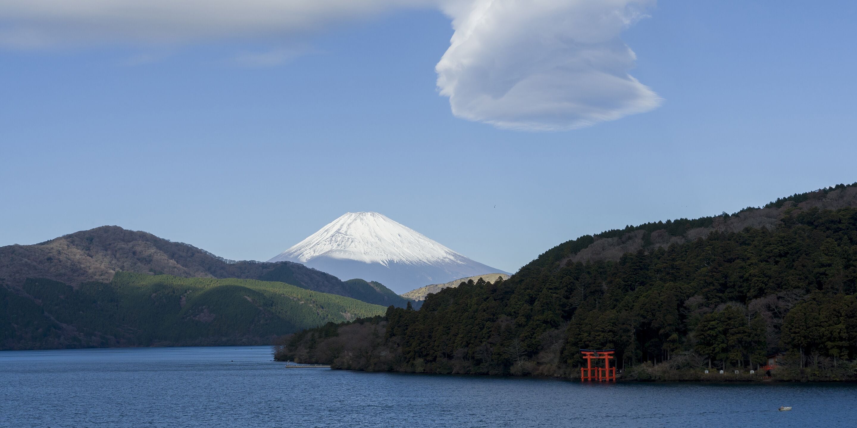 A mountain with a snow-capped peak, a body of water, and a red torii gate on the shore.