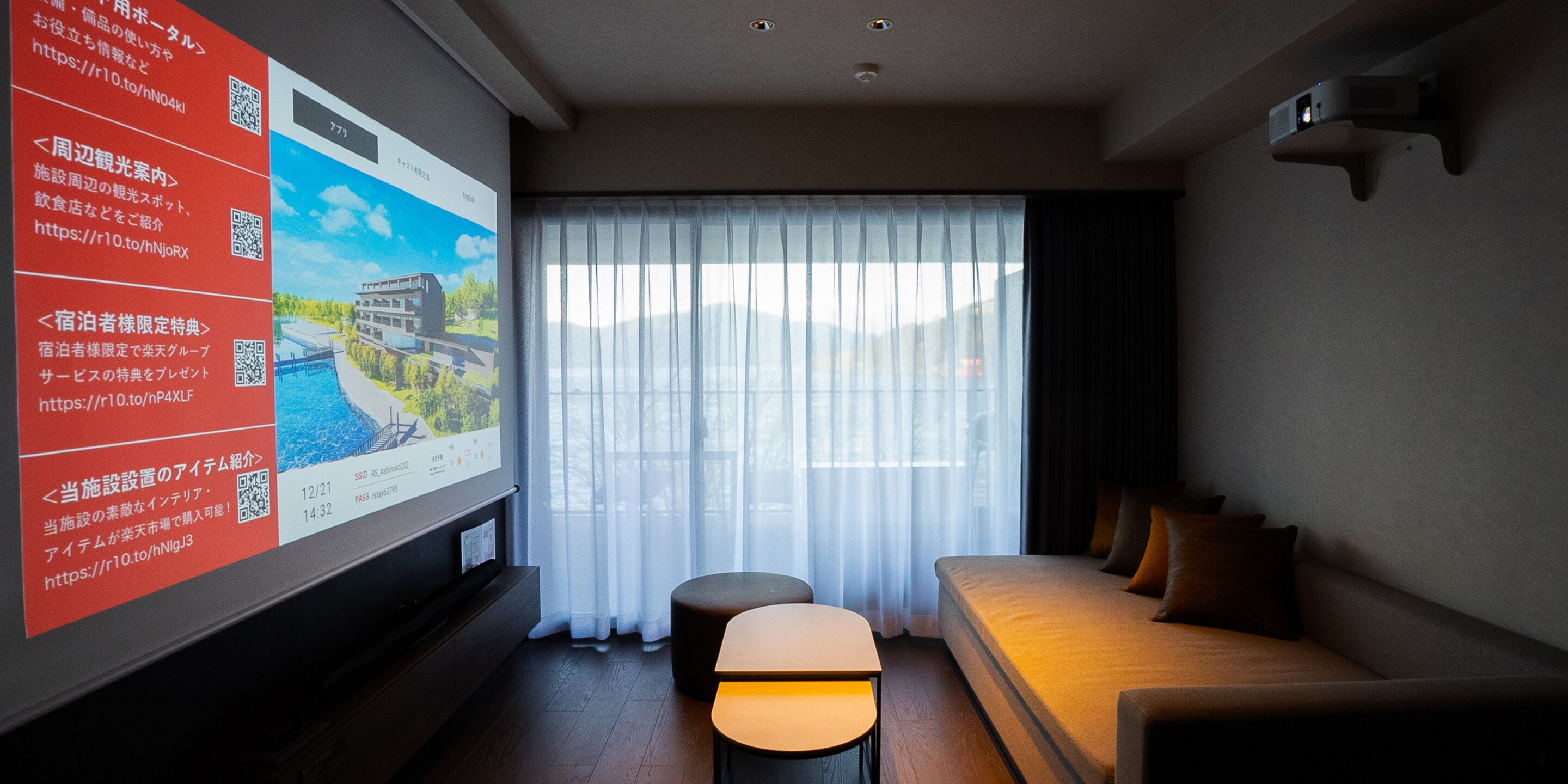 A hotel room with a large screen displaying information, a sofa, a small table, and a window with sheer curtains.