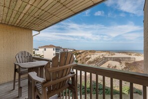 A wooden deck with two Adirondack chairs and a table, overlooking a beach with a wooden boardwalk and houses in the background.