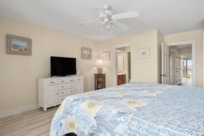 Une chambre à coucher avec un lit, un téléviseur posé sur une commode, un ventilateur de plafond et une porte donnant sur une autre pièce.