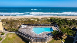 Piscina en la azotea con terraza de madera, rodeada por una valla de madera, con vista a una playa y al océano.
