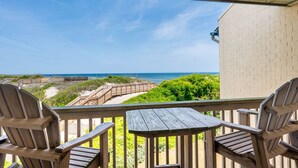 Una terraza de madera con dos sillas Adirondack y una mesa, con vista a una playa que tiene un paseo marítimo de madera y un edificio de ladrillo.