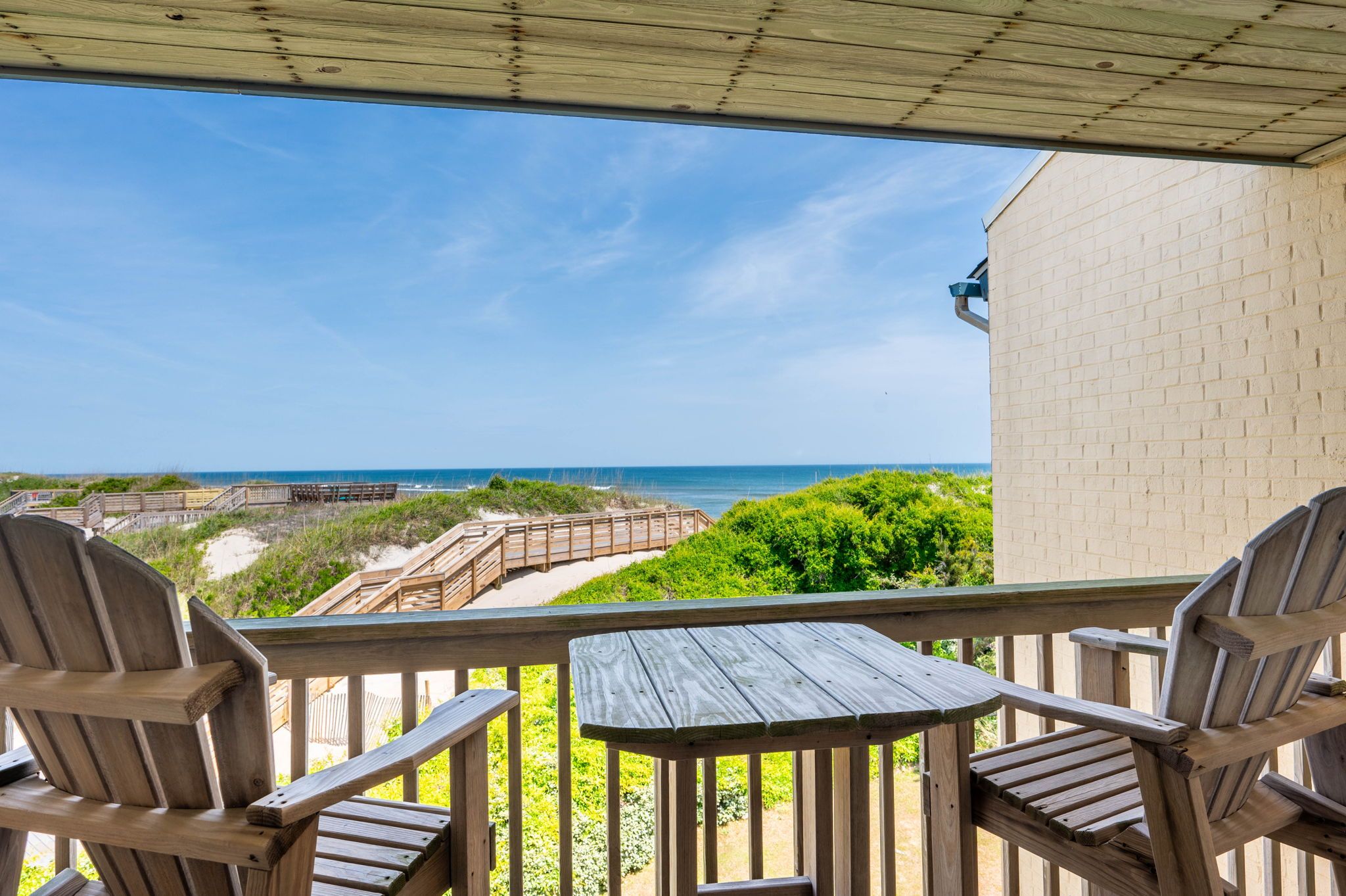 Una terraza de madera con dos sillas Adirondack y una mesa, con vista a una playa que tiene un paseo marítimo de madera y un edificio de ladrillo.