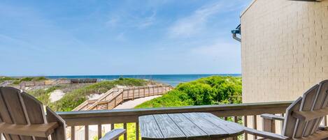 Una terraza de madera con dos sillas Adirondack y una mesa, con vista a una playa que tiene un paseo marítimo de madera y un edificio de ladrillo.