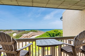 A wooden deck with two Adirondack chairs and a table, overlooking a beach with a wooden boardwalk and a brick building.