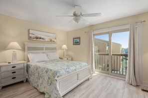 A bedroom with a bed, two bedside tables with lamps, a ceiling fan, a large window with curtains, and a balcony.