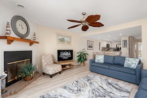 A living room with a fireplace, a television, a blue sofa, a ceiling fan, and a kitchen in the background.