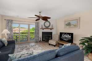 Una sala de estar amplia con un gran ventanal, ventilador de techo, chimenea y un televisor que muestra 'Coastal Carolina Vacations and Sales'.