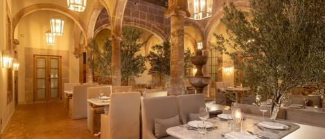 Un comedor interior con techos abovedados, paredes de piedra y olivos. Las mesas están dispuestas con platos, vasos y cubiertos.