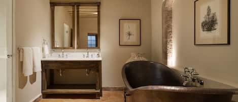 A bathroom with a stone-tiled floor, a wooden vanity with a large mirror, a freestanding bathtub, and framed artwork on the walls.