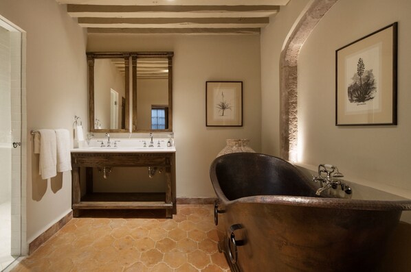 A bathroom with a stone-tiled floor, a wooden vanity with a large mirror, a freestanding bathtub, and framed artwork on the walls. - Casa Alondra (Morelia)