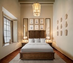 Un dormitorio con una cama grande, dos mesitas de noche con lámparas, un cabecero de madera, un ventanal con cortinas y jarrones en la pared.