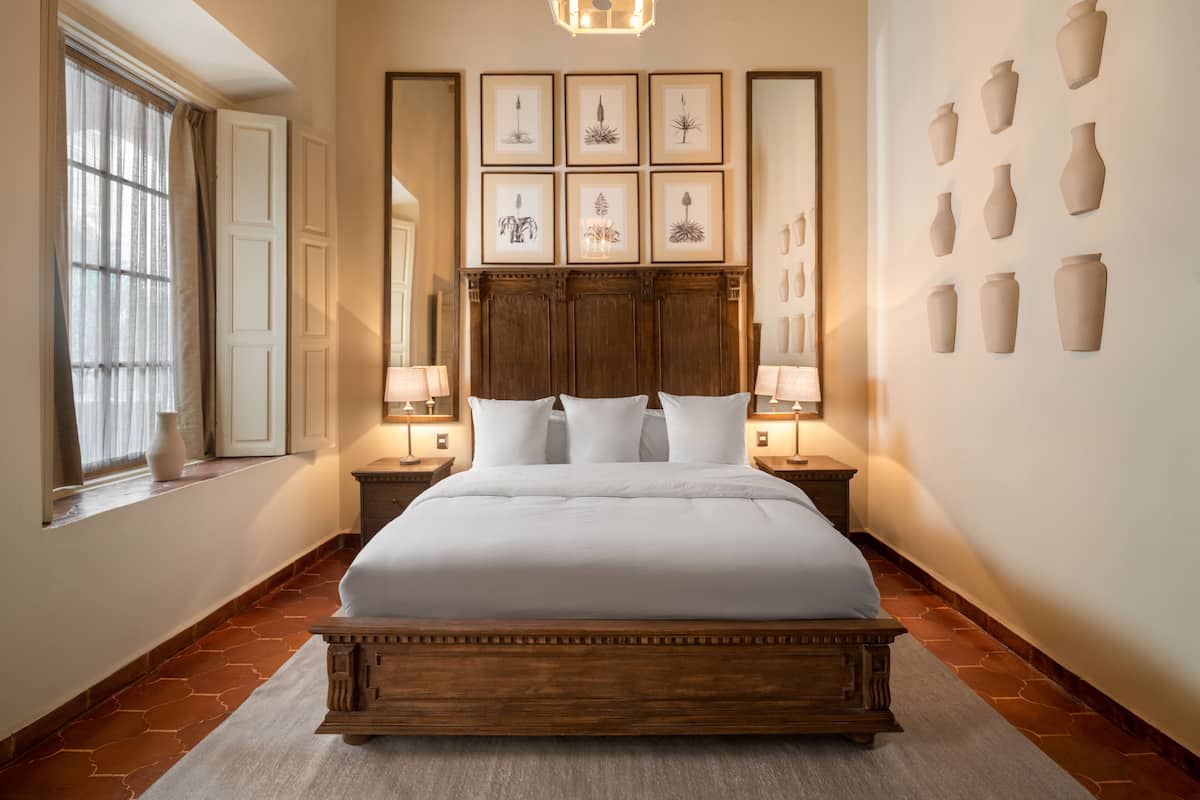 A bedroom with a large bed, two bedside tables with lamps, a chandelier, a large mirror, and framed botanical prints on the wall.