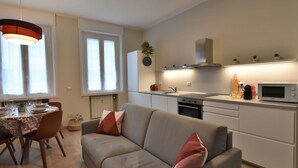Een moderne keuken met witte kastjes, een inbouwoven en een magnetron. Er is een zithoek met een tafel voor twee personen, een bank met kussens en een hanglamp boven de tafel.