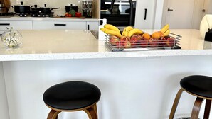 Une cuisine moderne avec des armoires blanches, une cuisinière en acier inoxydable et un panier de fruits posé sur le comptoir. On y trouve également deux tabourets en bois avec des assises noires.