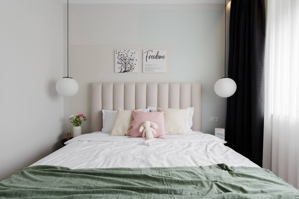 A neatly made bed with a green blanket, a white headboard, and decorative pillows. There are two wall-mounted light fixtures and a framed artwork above the bed.