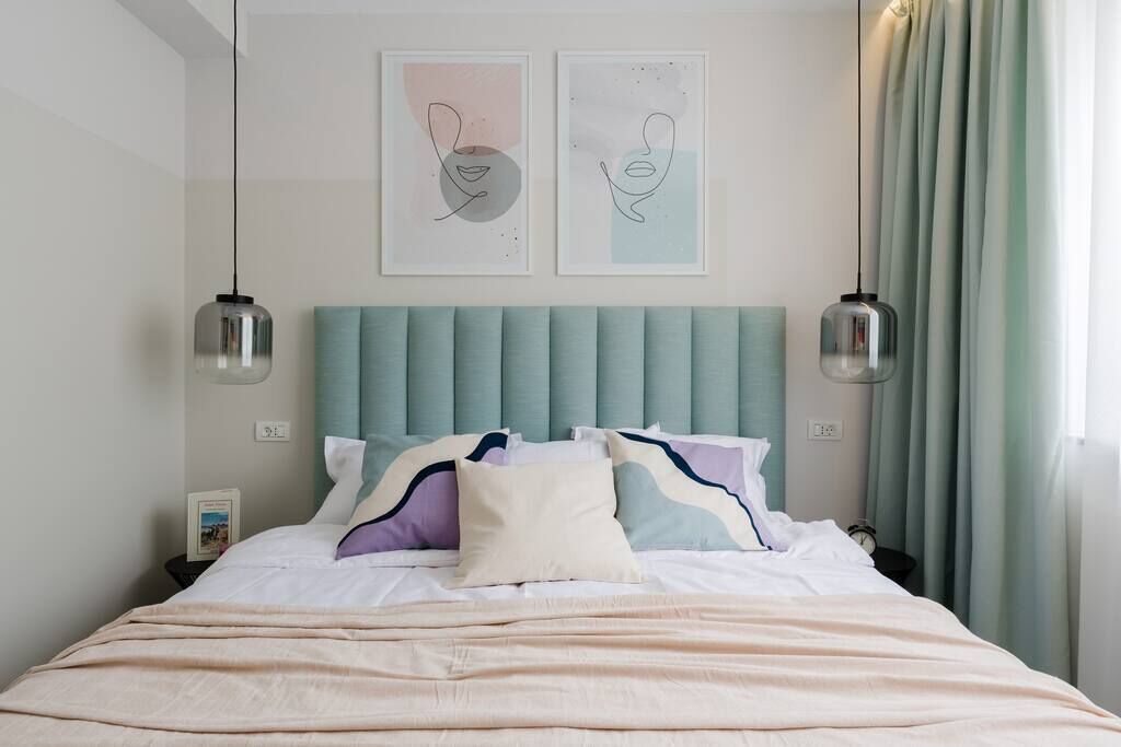 A neatly made bed with a light-colored duvet and pillows, a teal upholstered headboard, and two wall-mounted pendant lights.