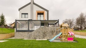 A modern house with a stone facade, a large glass window, and a wooden play structure with a slide.