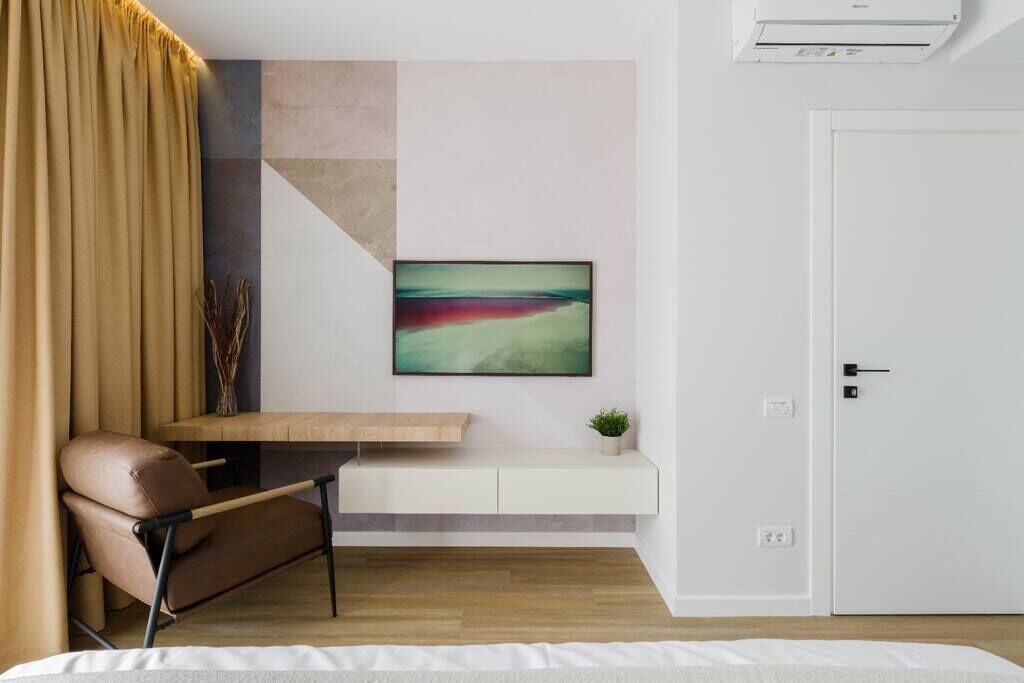 A modern room with a wooden desk, a brown leather chair, a white bedside table, a wall-mounted air conditioner, and a door.