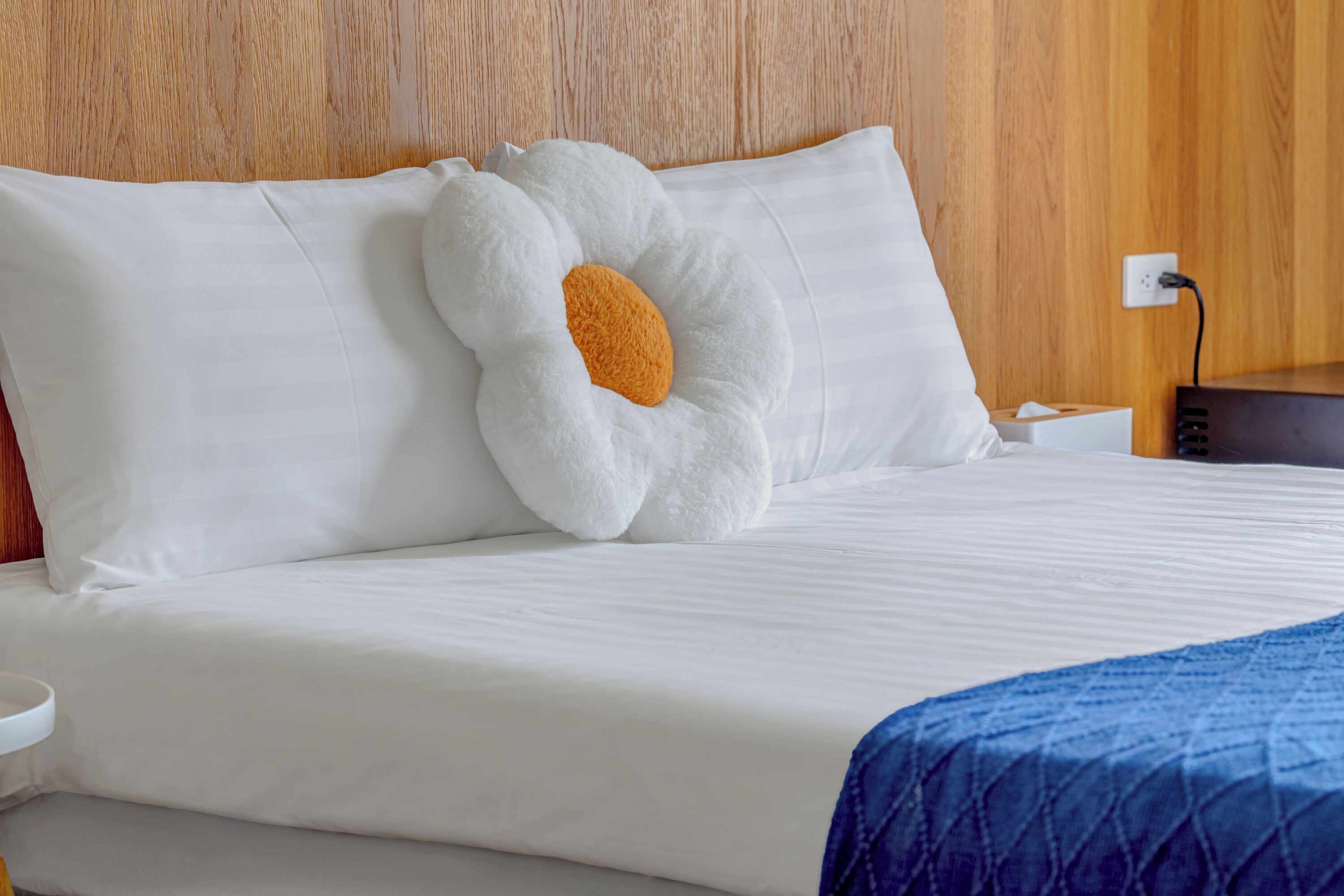 A neatly made bed with white linens, a blue blanket, and a unique white pillow with an orange center.