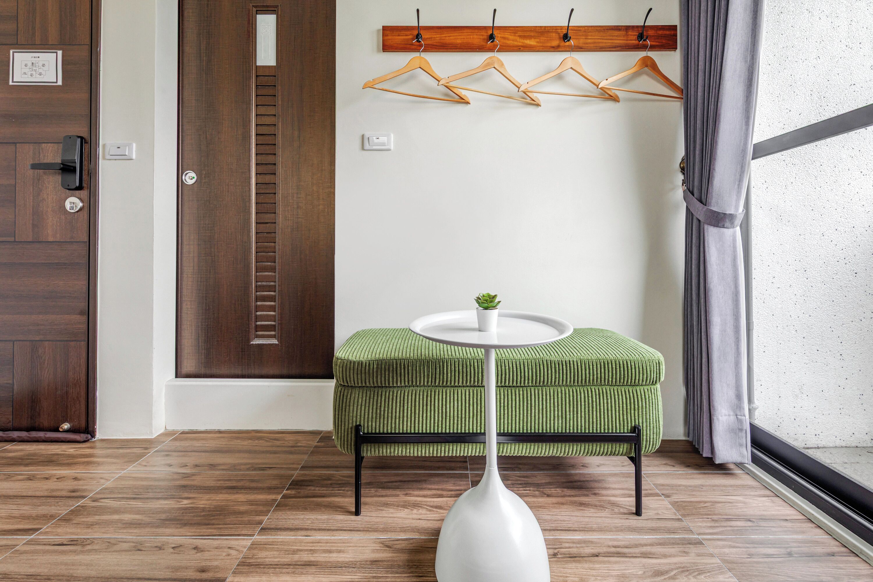 A room with a wooden door, a green upholstered bench, a white vase, and wooden hangers on a wall.