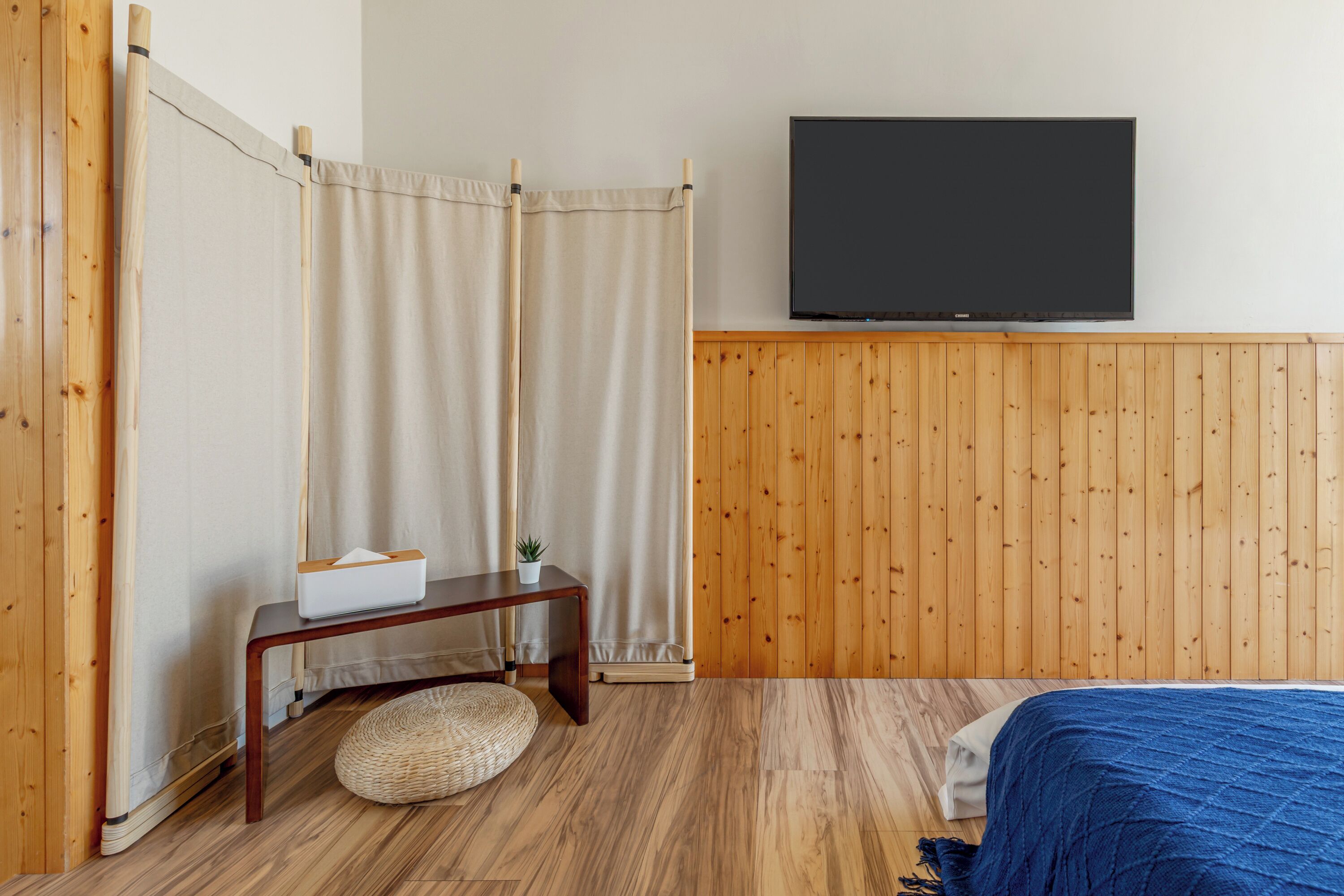 A bedroom with a wooden floor, a bed with a blue blanket, a TV on the wall, a bedside table with a tissue box, and a curtain.