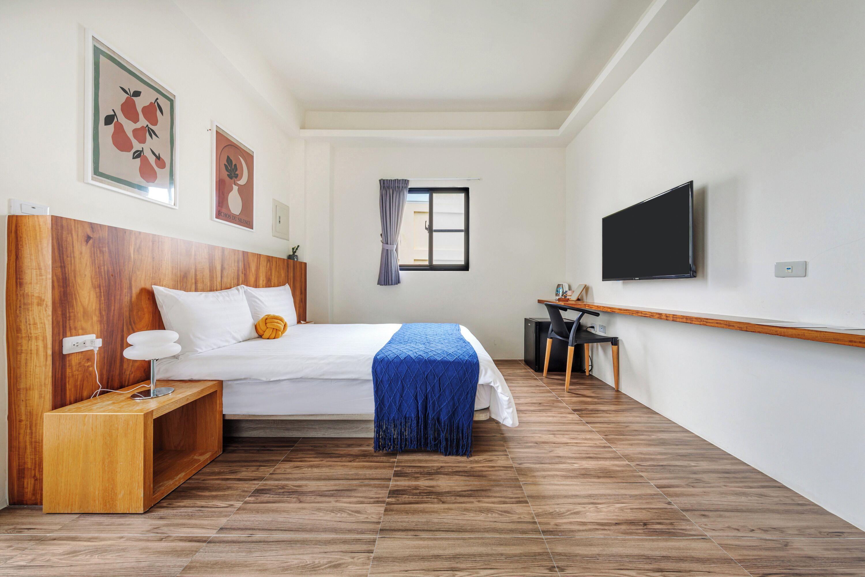 A bedroom with a wooden headboard, a bed with white bedding, a blue throw, a desk with a chair, and a TV on the wall.