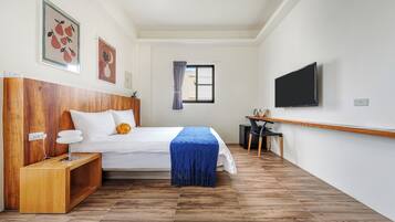 A bedroom with a wooden headboard, a bed with white bedding, a blue throw, a desk with a chair, and a TV on the wall.