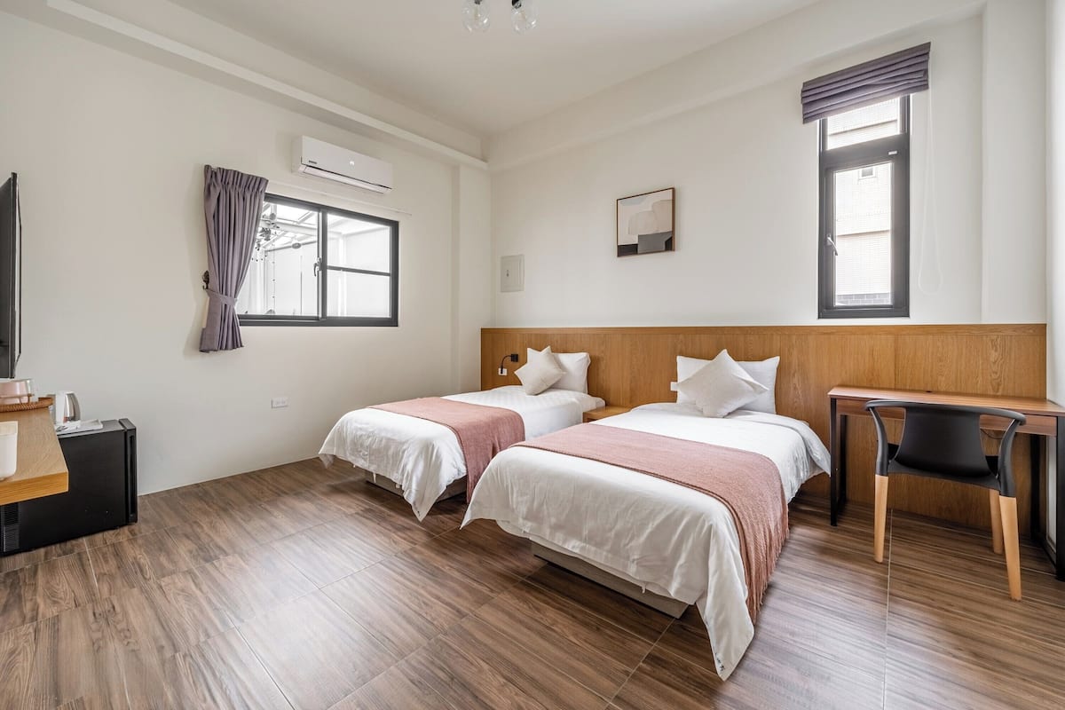 A hotel room with two beds, a desk, a chair, a TV, and an air conditioning unit.