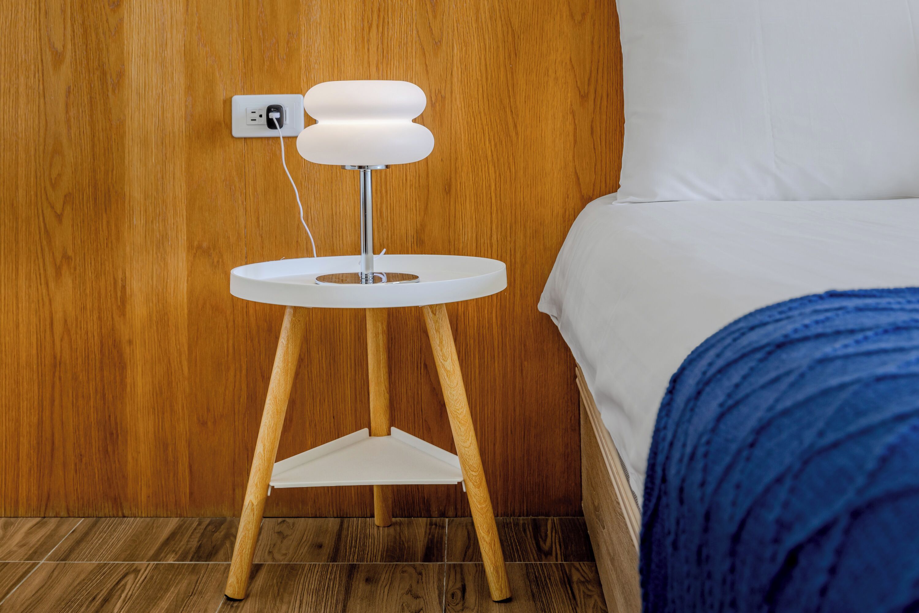 A wooden wall, a white bedside table with a lamp, a bed with white bedding, and a blue blanket.