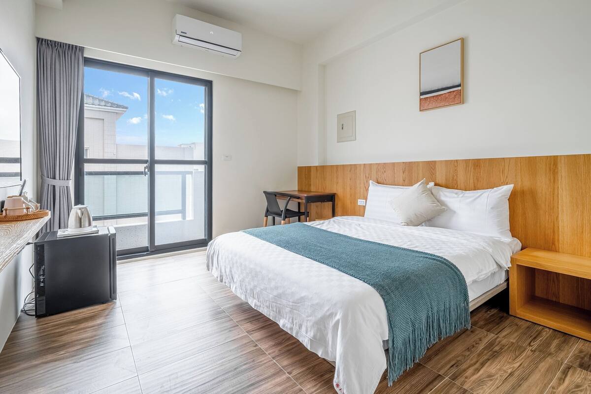 A hotel room with a large bed, a wooden headboard, a small table, a chair, a desk, and a window with a view of buildings.