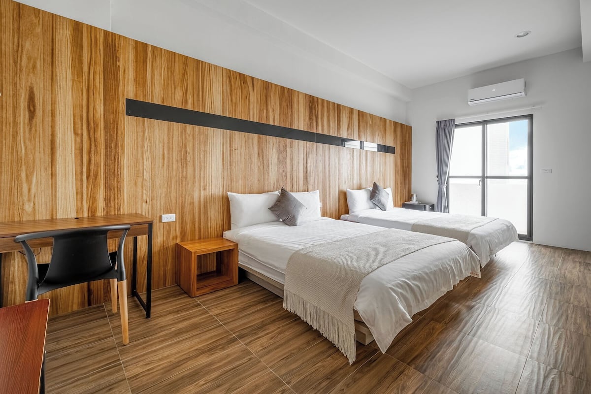 A modern hotel room with a wooden accent wall, a desk, two beds, and a large window.