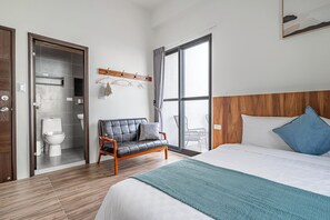 Ein modernes Hotelzimmer mit einem großen Bett, einem hölzernen Kopfteil, einer Sitzecke mit einer Ledersofa und Blick durch eine Schiebetür sowie einem kleinen Badezimmer mit Toilette und Waschbecken.