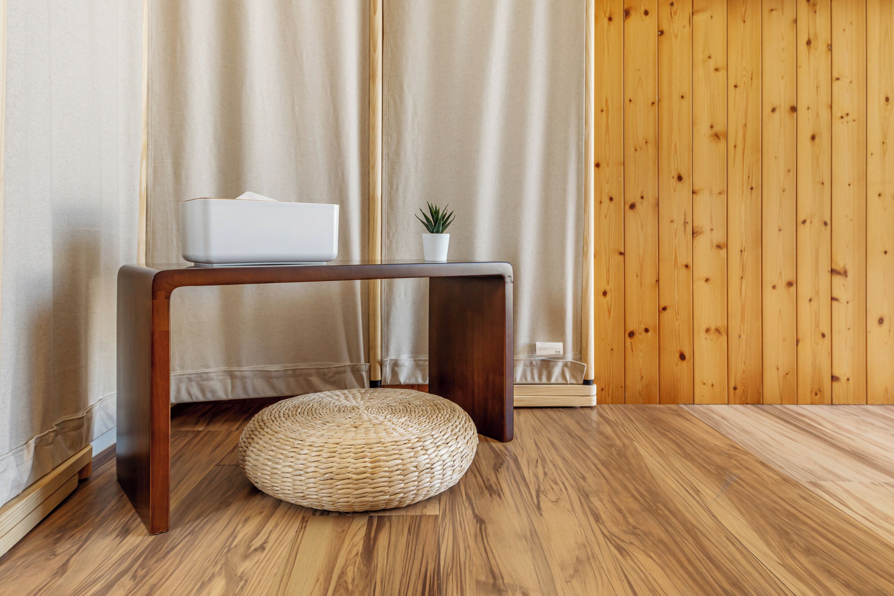 Un plancher en bois, un pouf en osier, un objet blanc posé sur une console en bois, et une petite plante en pot.