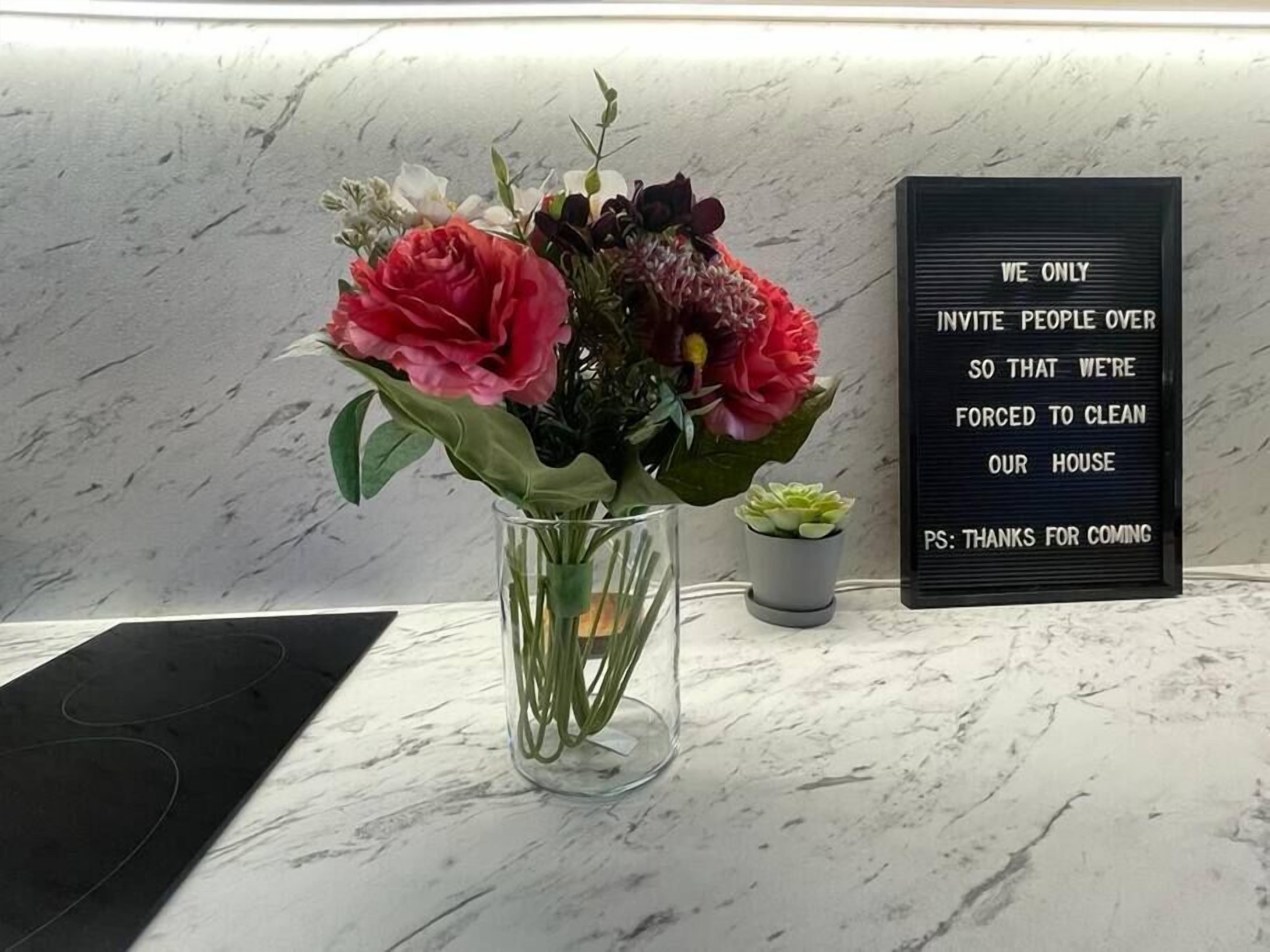 Una encimera de mármol con un florero transparente con flores y una pequeña planta en maceta. En el fondo se ve un letrero negro con letras blancas.