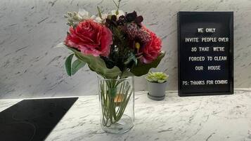 Una encimera de mármol con un florero transparente con flores y una pequeña planta en maceta. En el fondo se ve un letrero negro con letras blancas.
