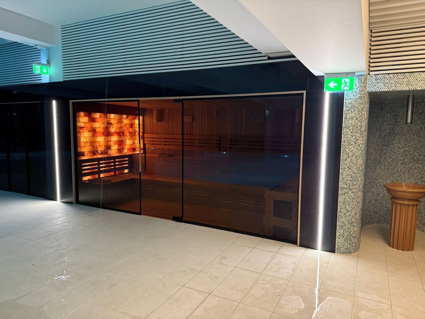 Un edificio moderno con entrada de cristal, un área de sauna visible con un banco de madera y un piso de baldosas.
