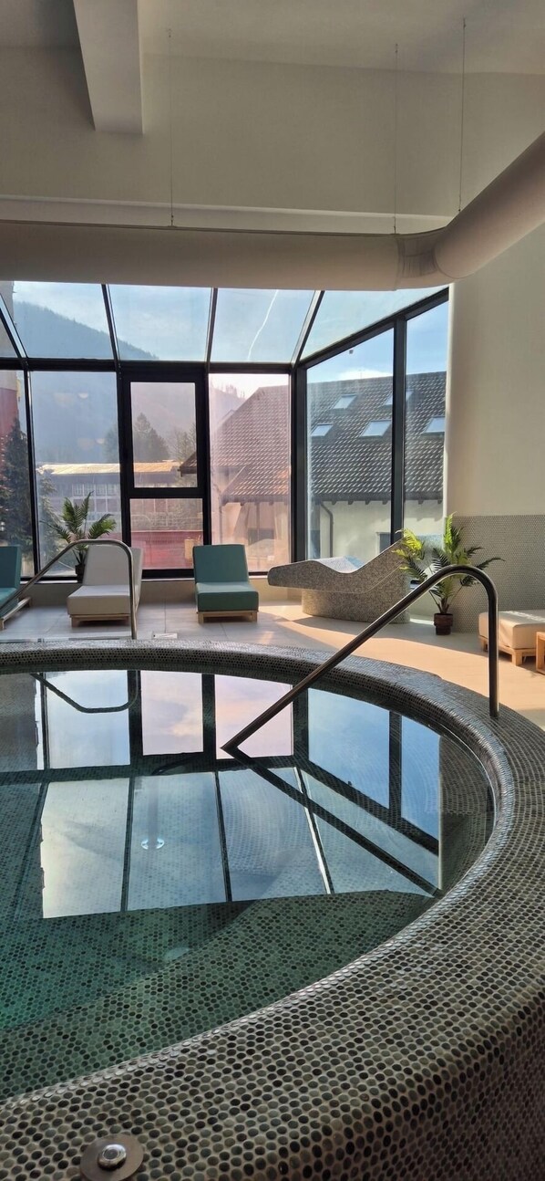 A modern indoor pool area with clear glass walls, seating, and a view of the outside.