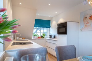 A modern kitchen with a sink, stove, and window with a blue valance. There is a dining area with a table and chairs. - Languste 2 - Traumhafte Ferienwohnung in Alt Westerland: Moderne Eleganz und Liebe zum Detail (Sylt)