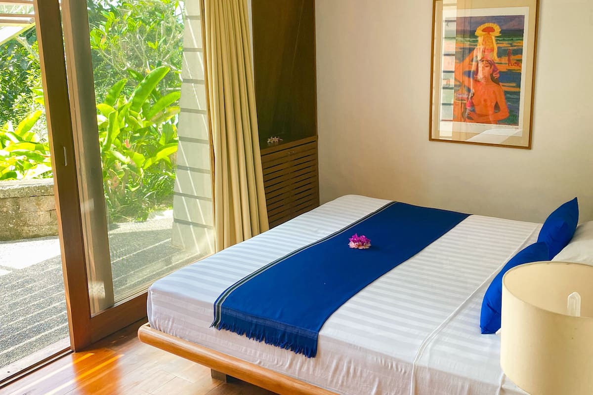 A bedroom with a bed, a nightstand, a lamp, a painting, and a sliding glass door leading to a balcony.
