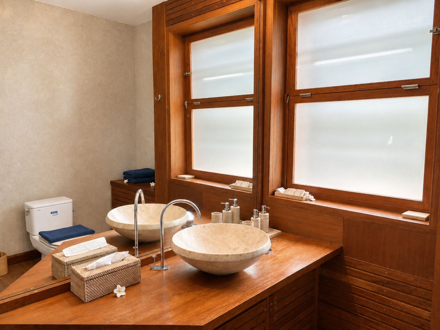 Un baño con dos lavamanos grandes y redondos color beige, un mueble de tocador de madera, un inodoro y una ventana con vidrio esmerilado.