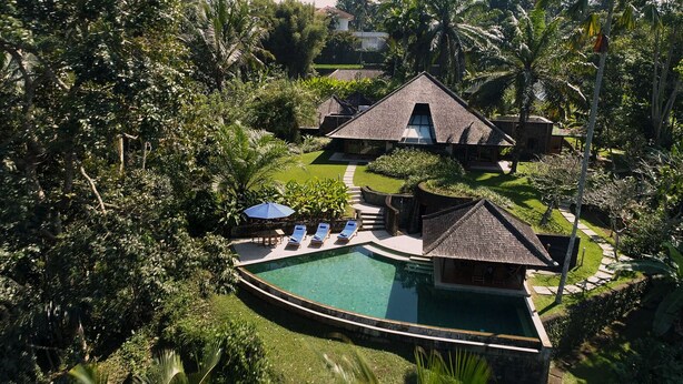 Un resort con piscina, sillones de descanso y un edificio con techo de paja, rodeado de exuberante vegetación.