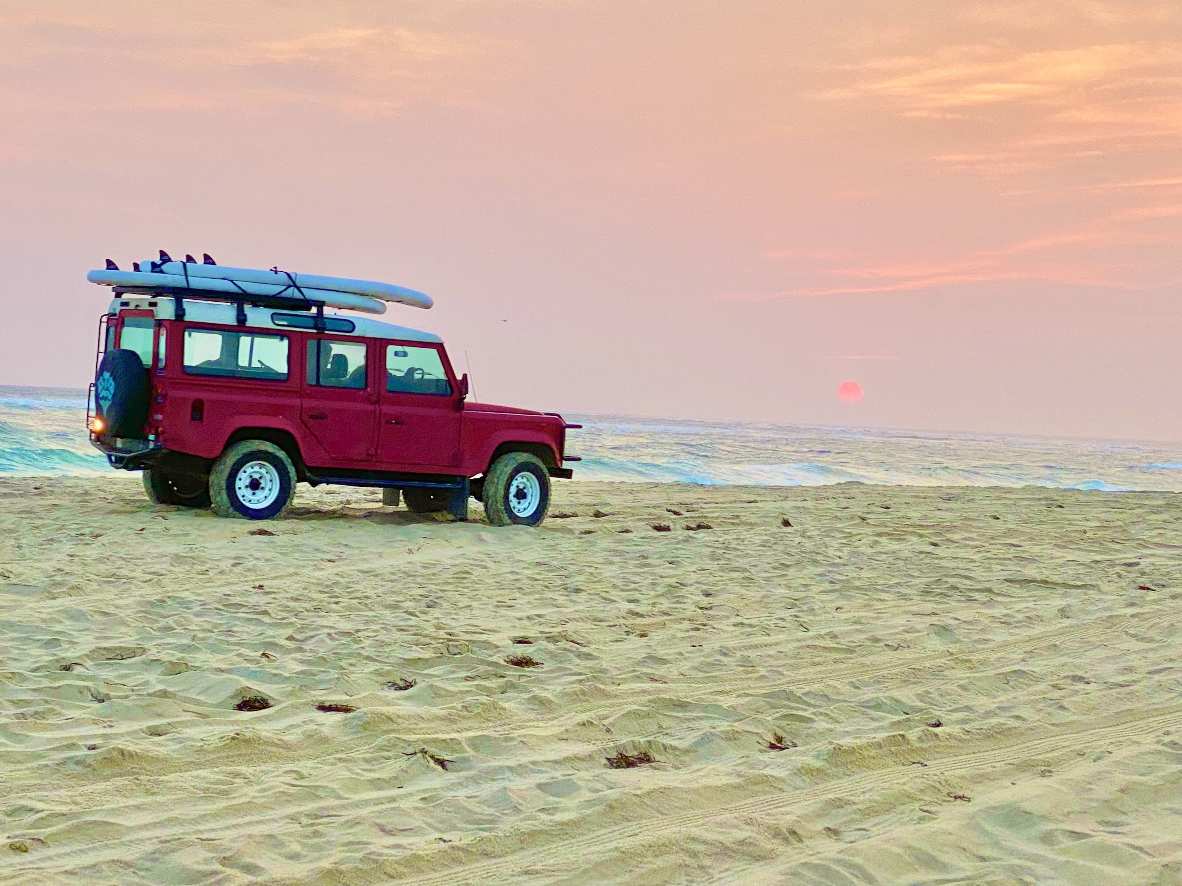 Un véhicule tout-terrain rouge est garé sur une plage de sable, avec des planches de surf sur son toit.