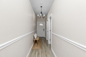 A narrow hallway with a bench, a door at the end, and a chandelier.