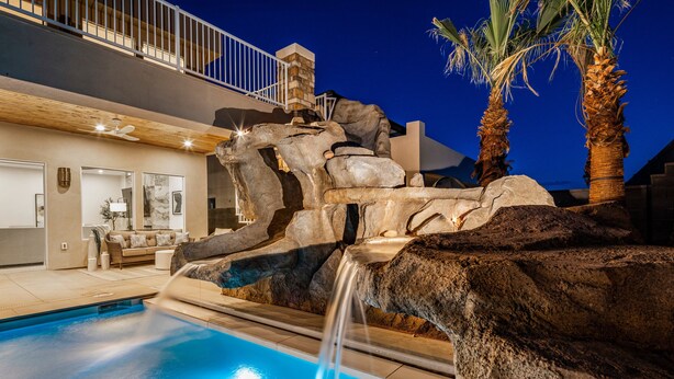 A swimming pool with a waterfall feature, surrounded by large rocks and palm trees. - Pecan Valley Resort| BRAND NEW Private Pool and Hot Tub w/ Slide (Hurricane)