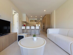 A modern living room with a flat-screen TV, a white sofa, a round coffee table, and a kitchen area with bar stools and a countertop.