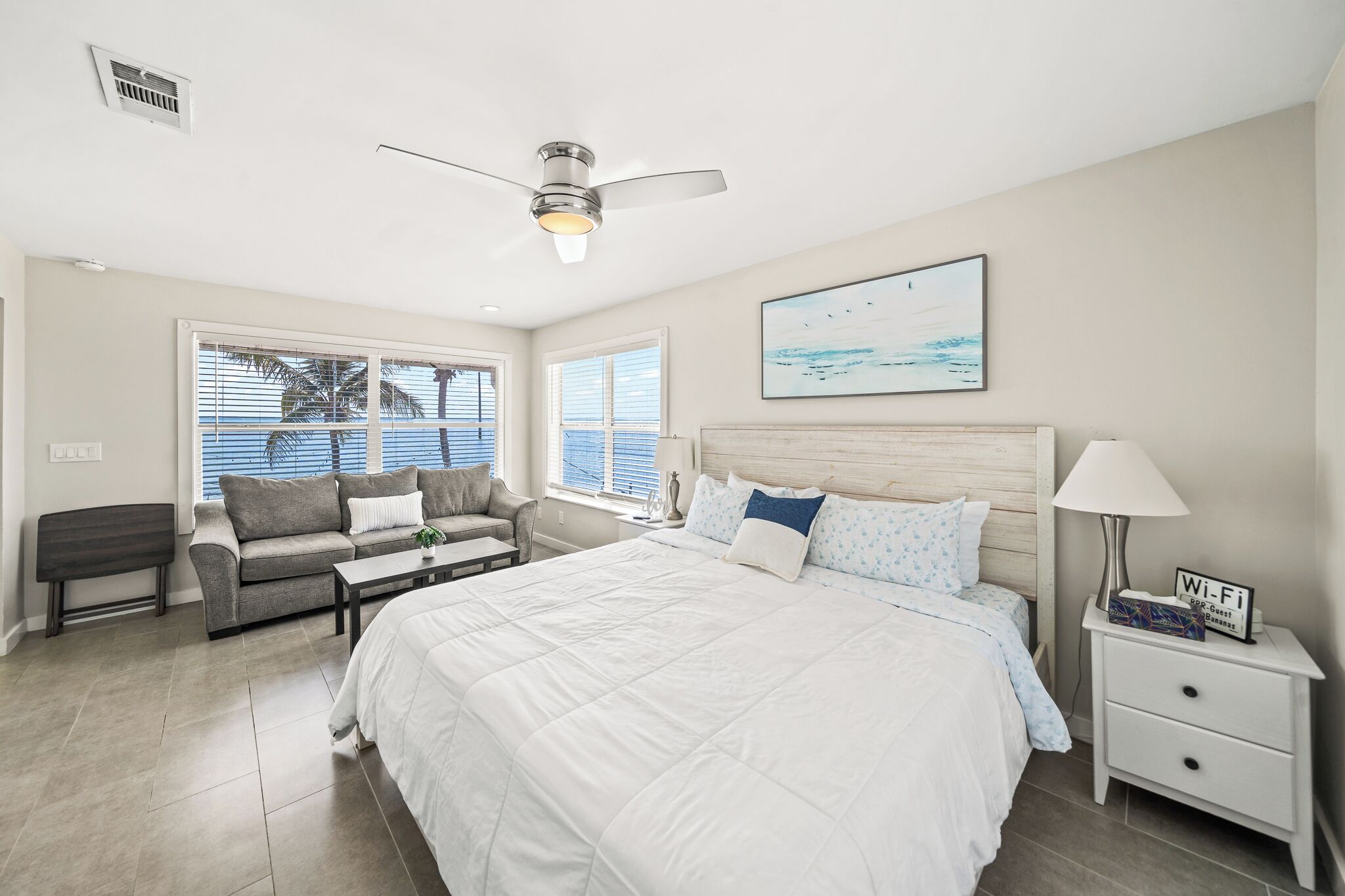 A bedroom with a large bed, a ceiling fan, a nightstand with a lamp, and a window with blinds.